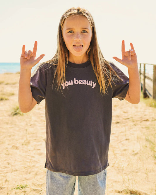 A young girl with long light brown hair stands on a sandy beach, making a peace sign with both hands and sticking her tongue out. She is wearing a faded black graphic t-shirt with the words 'you beauty' printed in white across the chest, paired with light wash blue jeans.
