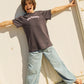 A person wearing an oversized dark charcoal t-shirt with the text 'you beauty' in white script, paired with light-wash wide-leg jeans, posing against a white wooden wall.